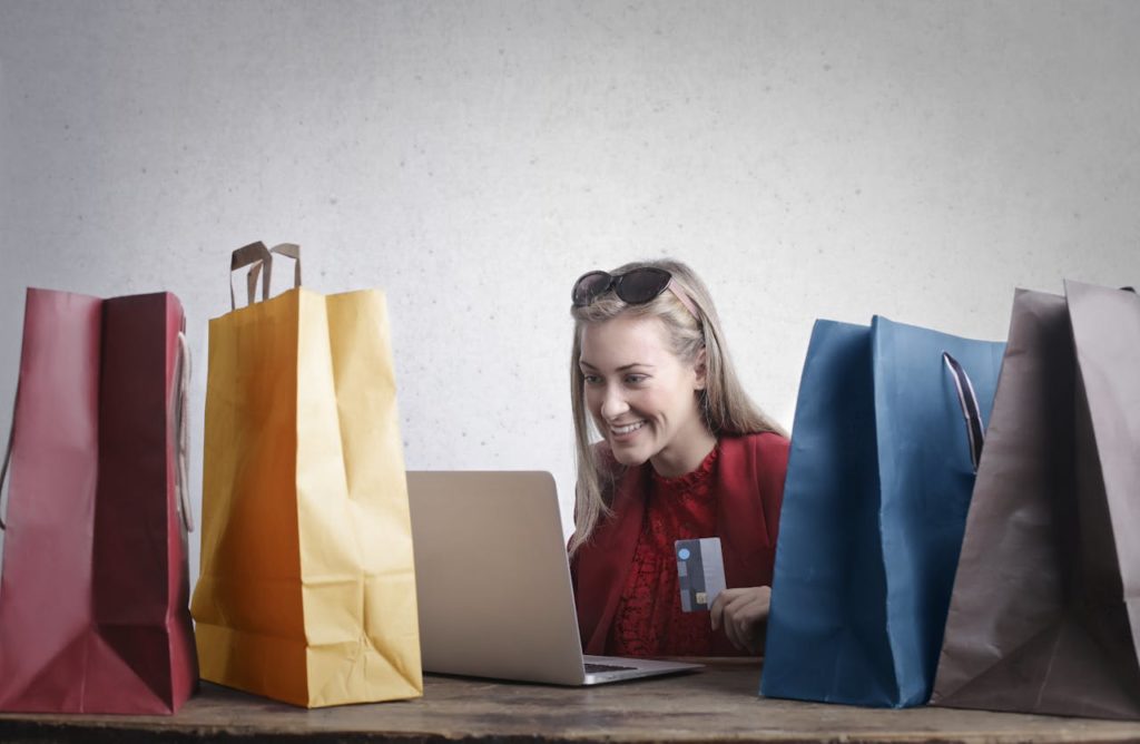 Jeune femme en train de passer des commandes depuis son ordinateur portable, entourée de sacs en papier de couleur.