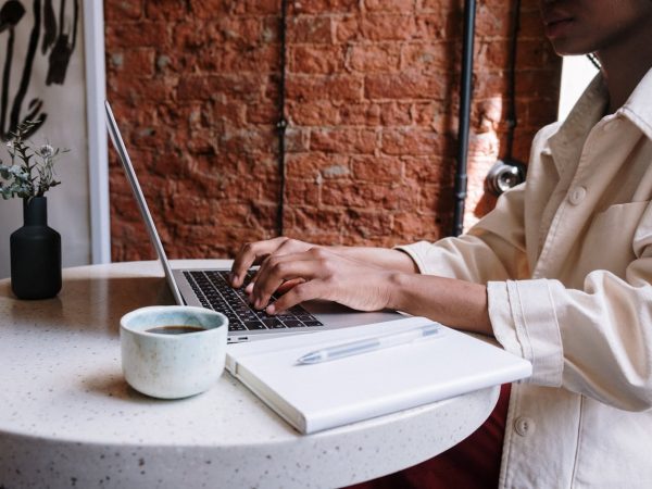 Jeune femme assise dans un café en train d'écrire à l'ordinateur avec un café et un carnet.